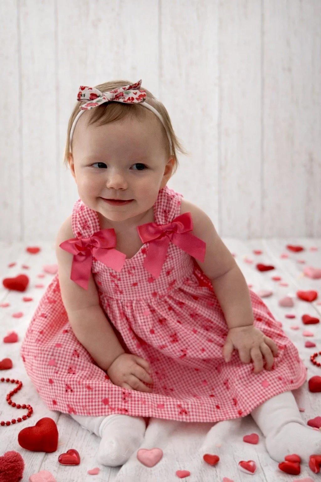 A young girl with a headband and a pink checkered dress sitting among Valentine's Day hearts and decorations on a white wooden floor, with a light wooden panel background.
