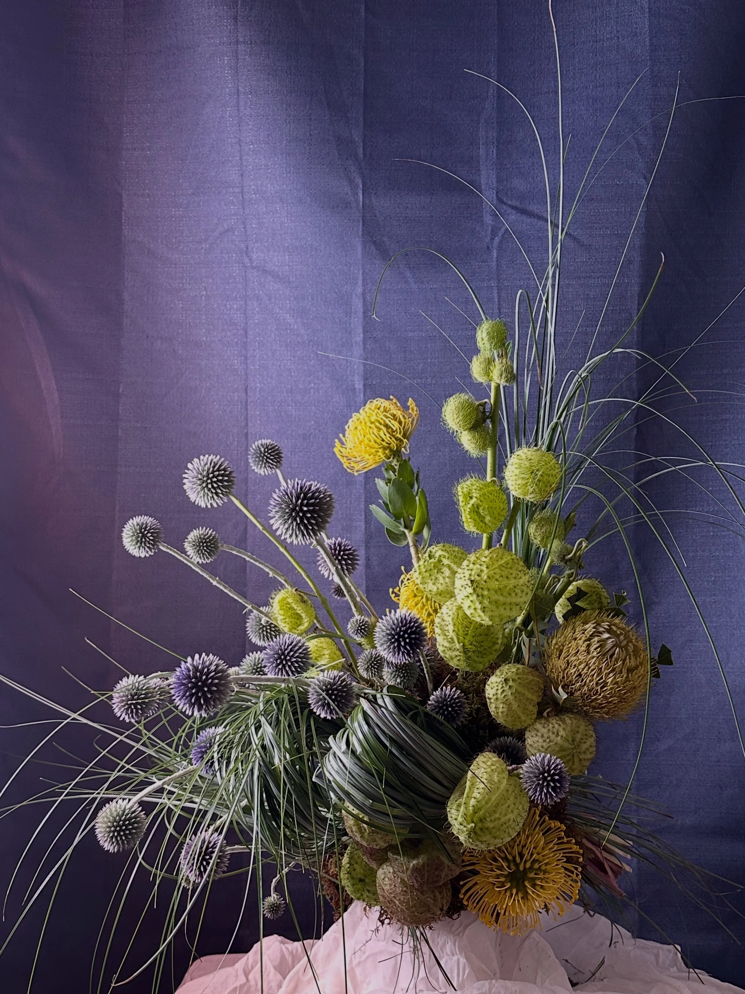 A tall bouquet of various flowers and grasses against a purple fabric background.