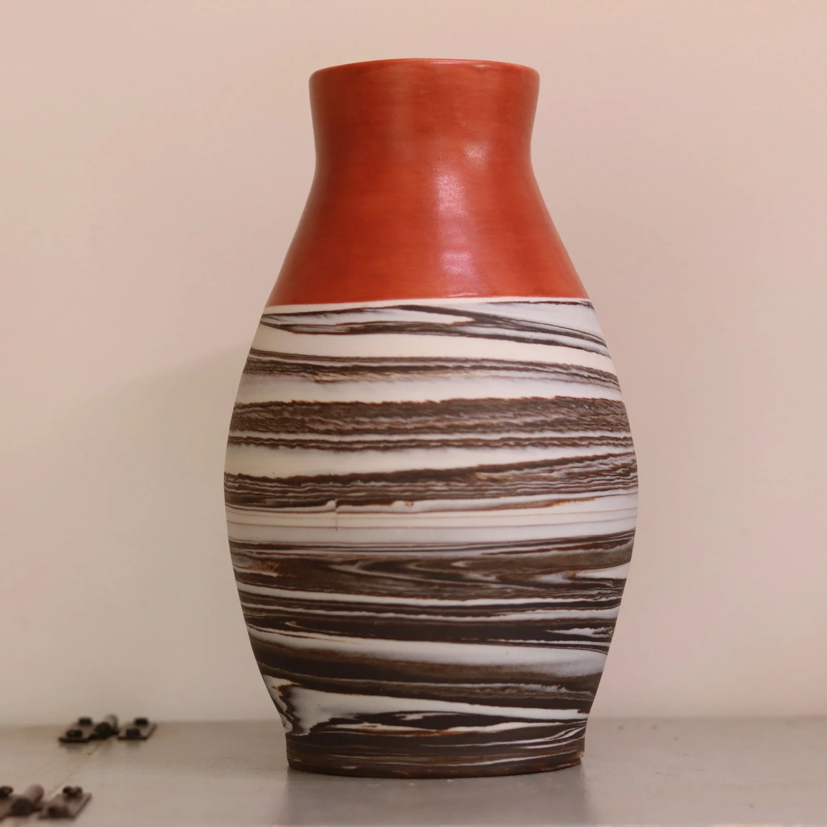 A ceramic vase with red upper part and black and white striped lower part, placed on a flat surface with some small objects or debris in the background.