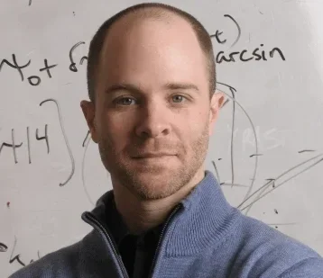 A man with short hair and a beard standing in front of a whiteboard with writing and diagrams.