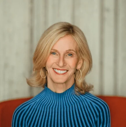 A woman with shoulder-length blonde hair wearing a blue ribbed turtleneck sweater, smiling, sitting in front of a light-colored wooden backdrop.