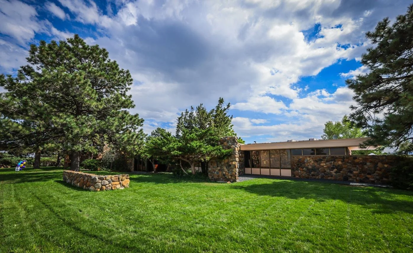 A mid-century modern house with a stone exterior surrounded by a lush, green lawn and tall trees on a partly cloudy day.