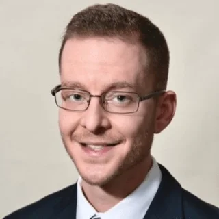A young man with short brown hair, glasses, and a light beard, smiling and wearing a dark blazer and a light shirt.
