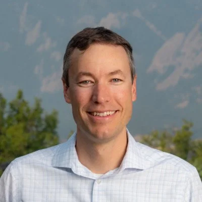 A man smiling outdoors in front of mountains and trees.
