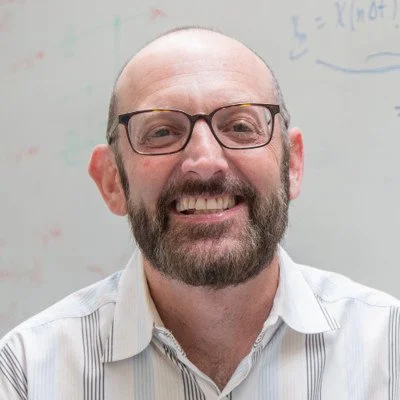 Smiling man with glasses and a beard in front of a whiteboard with writing in the background.