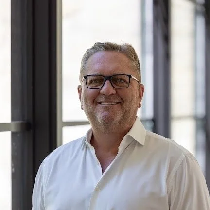 A smiling man with glasses and light-colored hair, wearing a white shirt, standing indoors near large windows.