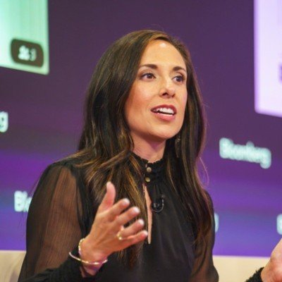 A woman with long dark hair speaking during a panel discussion, wearing a black sheer-sleeve top, with a purple background and partial view of a monitor or screen.
