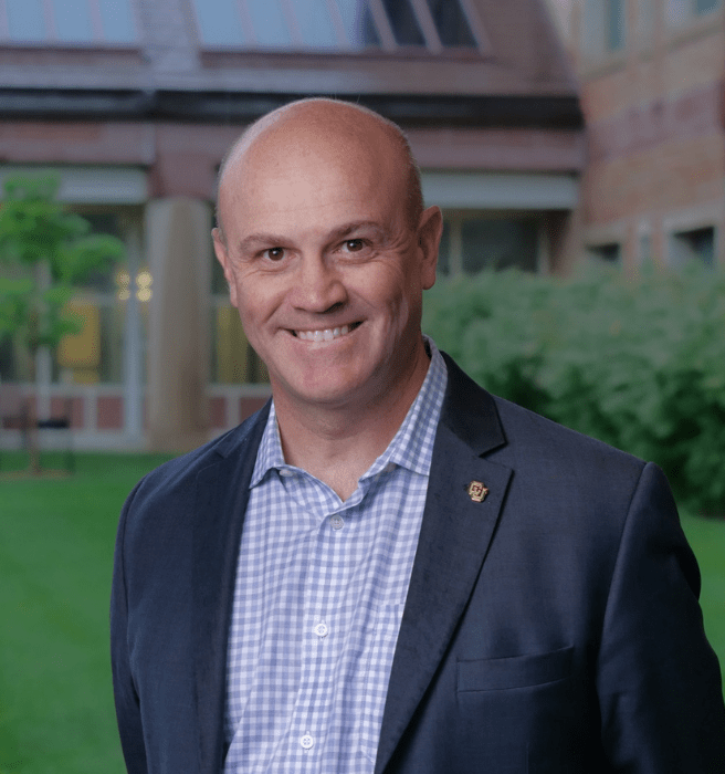 Smiling man in a blue suit and light blue checkered shirt outdoors with a building and greenery in the background.