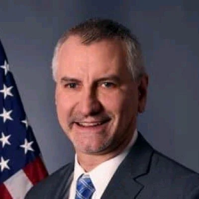 Portrait of a man in a suit and tie, standing in front of an American flag.