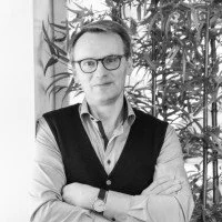 A man with glasses wearing a sweater vest over a collared shirt, standing with his arms crossed, in front of a background with leafy plants.