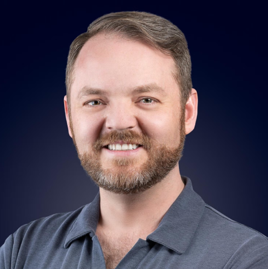 A smiling man with a beard and short brown hair, wearing a gray collared shirt, against a dark blue background.