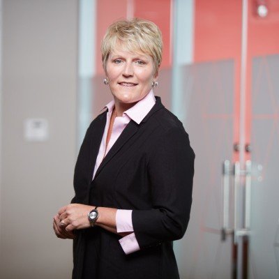 A professional woman with short blonde hair wearing a black blazer and pink shirt standing in an office hallway with red and gray walls and glass doors.