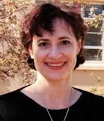 Close-up of a woman with short curly hair smiling outdoors, with trees and a building in the background.