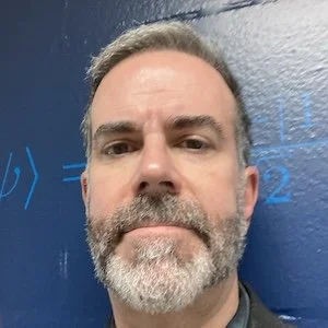Close-up of a man with gray hair and beard taking a selfie in front of a blue wall with some writing visible.