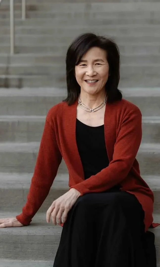 A woman with short black hair, wearing a red cardigan over a black top and black skirt, sitting on concrete stairs outdoors, smiling at the camera.