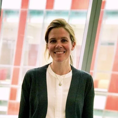 A woman smiling in front of large windows with a modern building background.