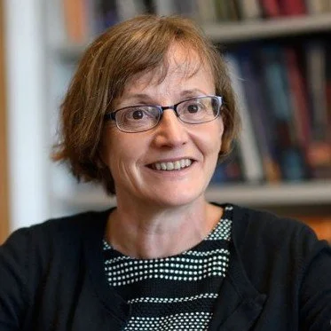 Portrait of a smiling woman with glasses and short brown hair, standing in front of bookshelves.