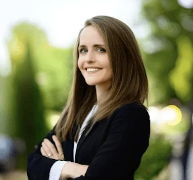 A young woman with long brown hair smiling outdoors, wearing a black blazer and white shirt.