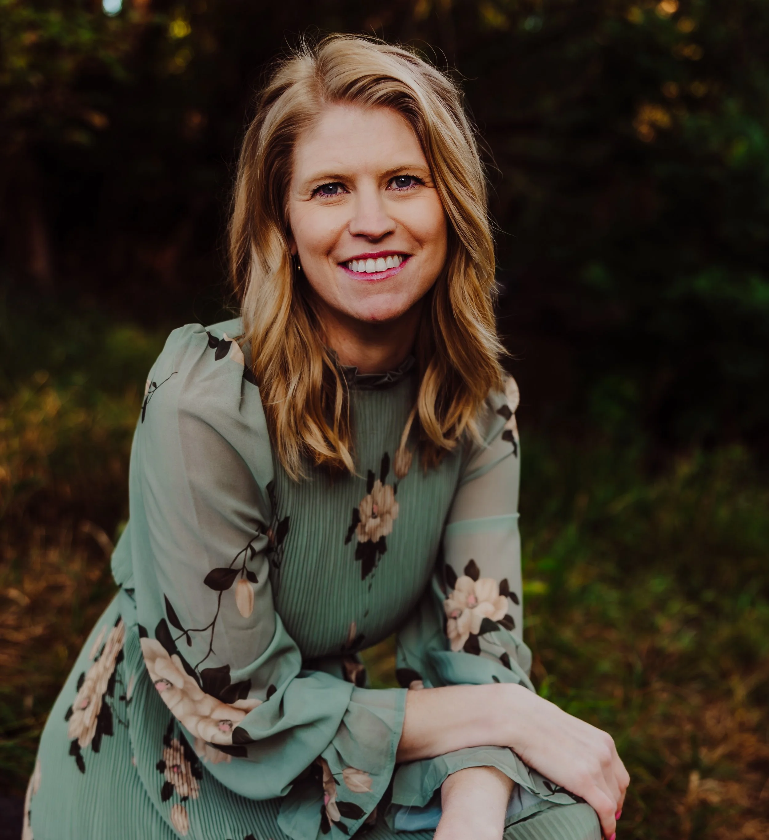 Woman in a green floral dress with ruffled sleeves, sitting outdoors and smiling at the camera.