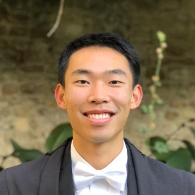 A young man wearing a tuxedo with a bowtie, smiling outdoors against a brick wall and greenery.