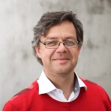A man with gray hair wearing glasses, a white collared shirt, and a red sweater, standing outdoors against a blurred background.