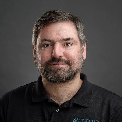 Portrait of a man with dark hair and beard wearing a black polo shirt against a gray background.