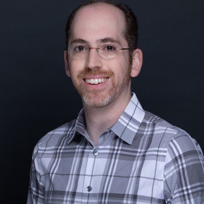 A man with glasses and a beard wearing a plaid shirt, smiling against a dark background.