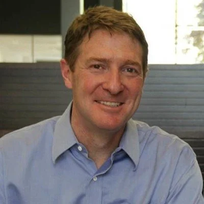 A man with brown hair and a blue collared shirt smiling at the camera in a modern office setting.
