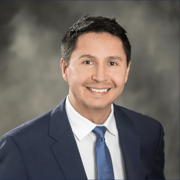 Professional headshot of a smiling man in a navy suit, white shirt, and blue tie against a gray blurred background.