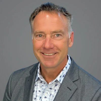 Portrait of a middle-aged man with short gray hair, smiling, wearing a gray blazer and a patterned white shirt against a plain gray background.