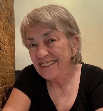 A smiling older woman with short gray hair, wearing a black shirt, sitting indoors near an exposed brick wall.