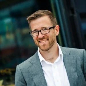 A smiling man with glasses in a gray suit and white shirt standing outdoors in front of a bus.