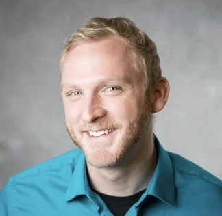 Headshot of a smiling man with blonde hair and a beard, wearing a teal shirt against a gray background.