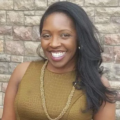 A smiling woman with long black hair, wearing a gold dress and a gold necklace, standing in front of a brick wall.