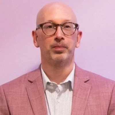 A bald man wearing glasses and a pink checkered blazer standing against a plain light purple background.