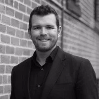 A man with short hair and a beard, wearing a dark blazer and shirt, smiling and standing outdoors against a brick wall.