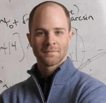 Portrait of a man with short hair and a beard, wearing a gray zip-up sweater, standing in front of a whiteboard with writing and diagrams.