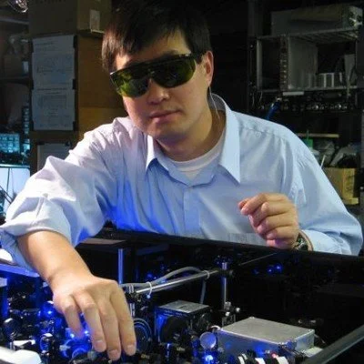 A man with dark hair wearing sunglasses and a white shirt working on machinery or equipment in a laboratory or technical setting.