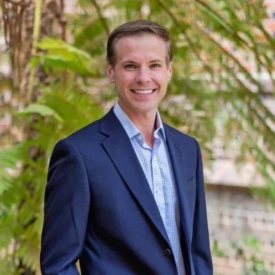 A man in a navy suit and light blue shirt smiling outdoors with green foliage in the background.
