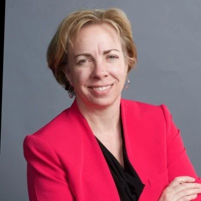 A woman with short blonde hair wearing a red blazer and black top, smiling with arms crossed against a gray background.