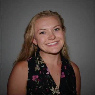 A young woman with blonde hair smiling, wearing a sleeveless black floral dress, against a gray background.