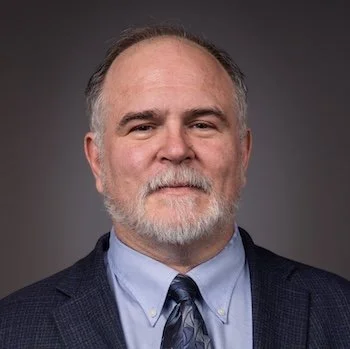 A middle-aged man with a gray beard and short gray hair, wearing a dark suit jacket, light blue shirt, and patterned tie, posing against a dark background.