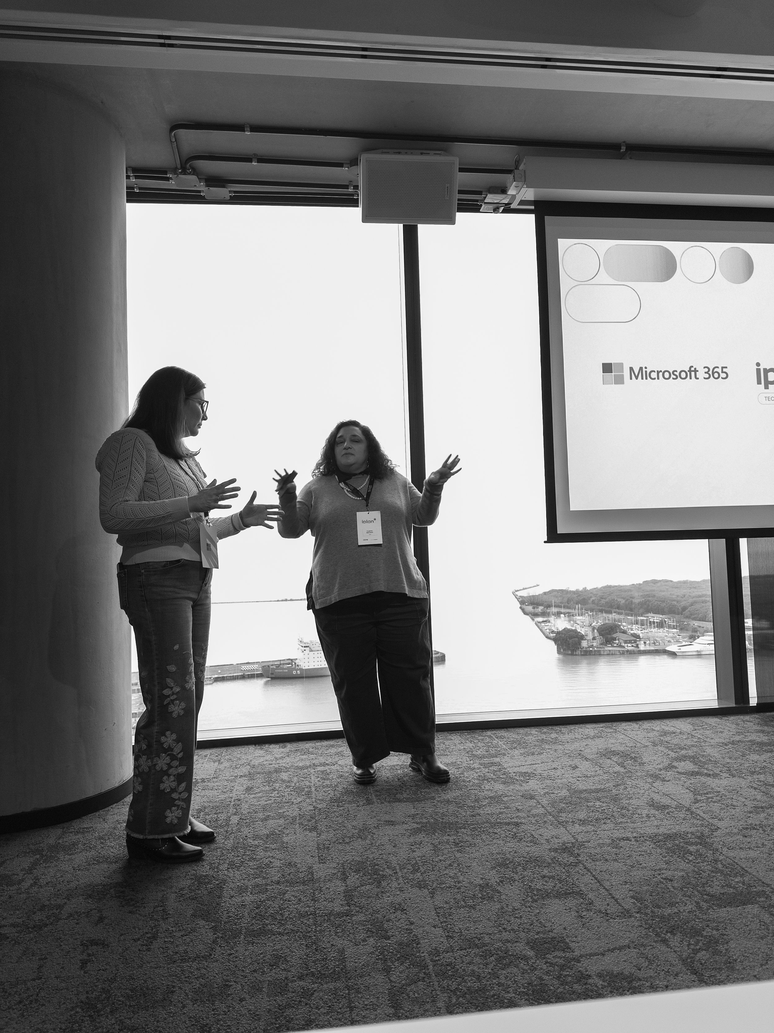Dos mujeres conversando en una sala con vista a un puerto y barcos, en una presentación de negocios con un proyector en la pared.