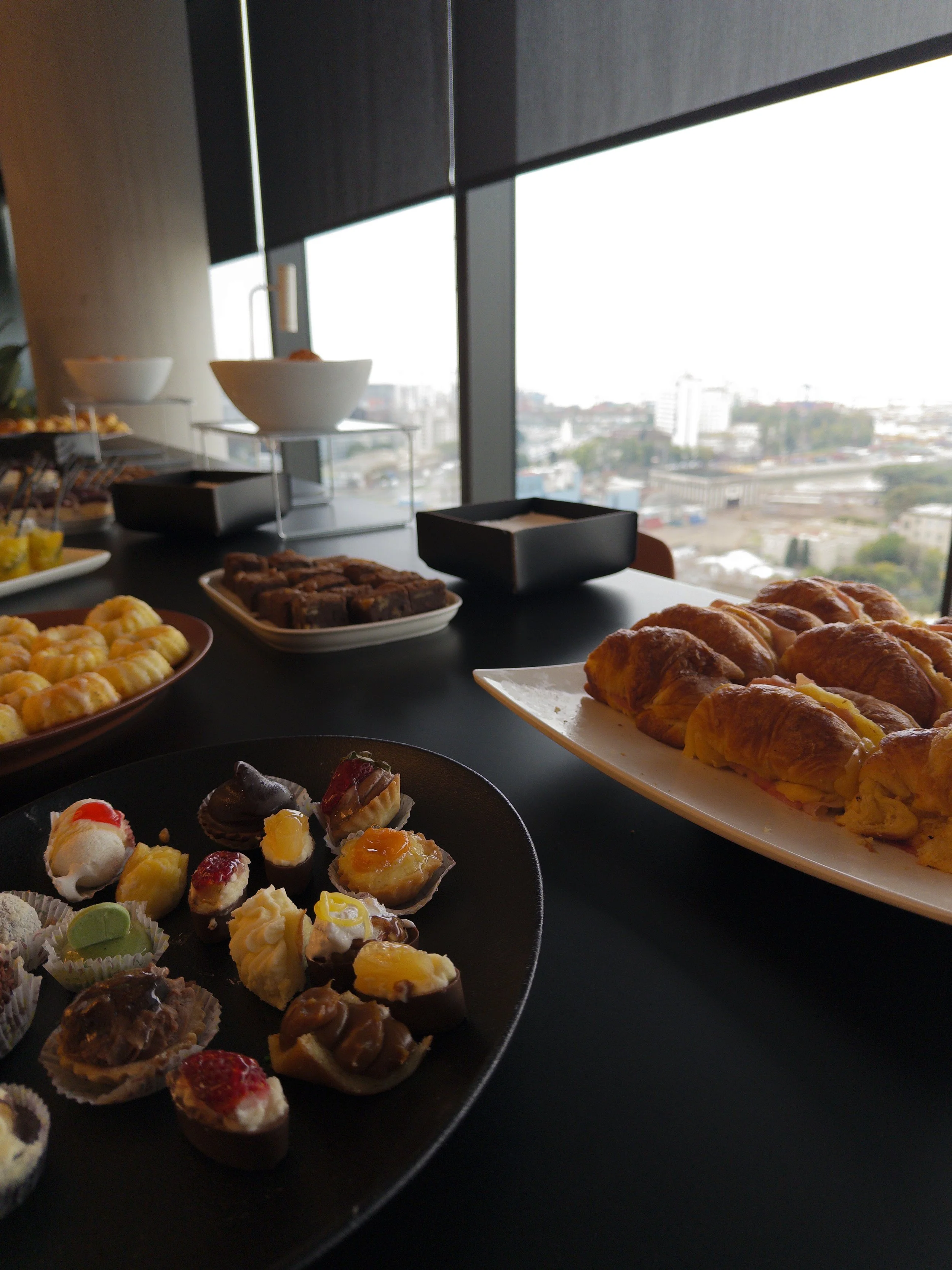 Variedad de postres y pasteles en una mesa cerca de una ventana grande con vista a la ciudad.