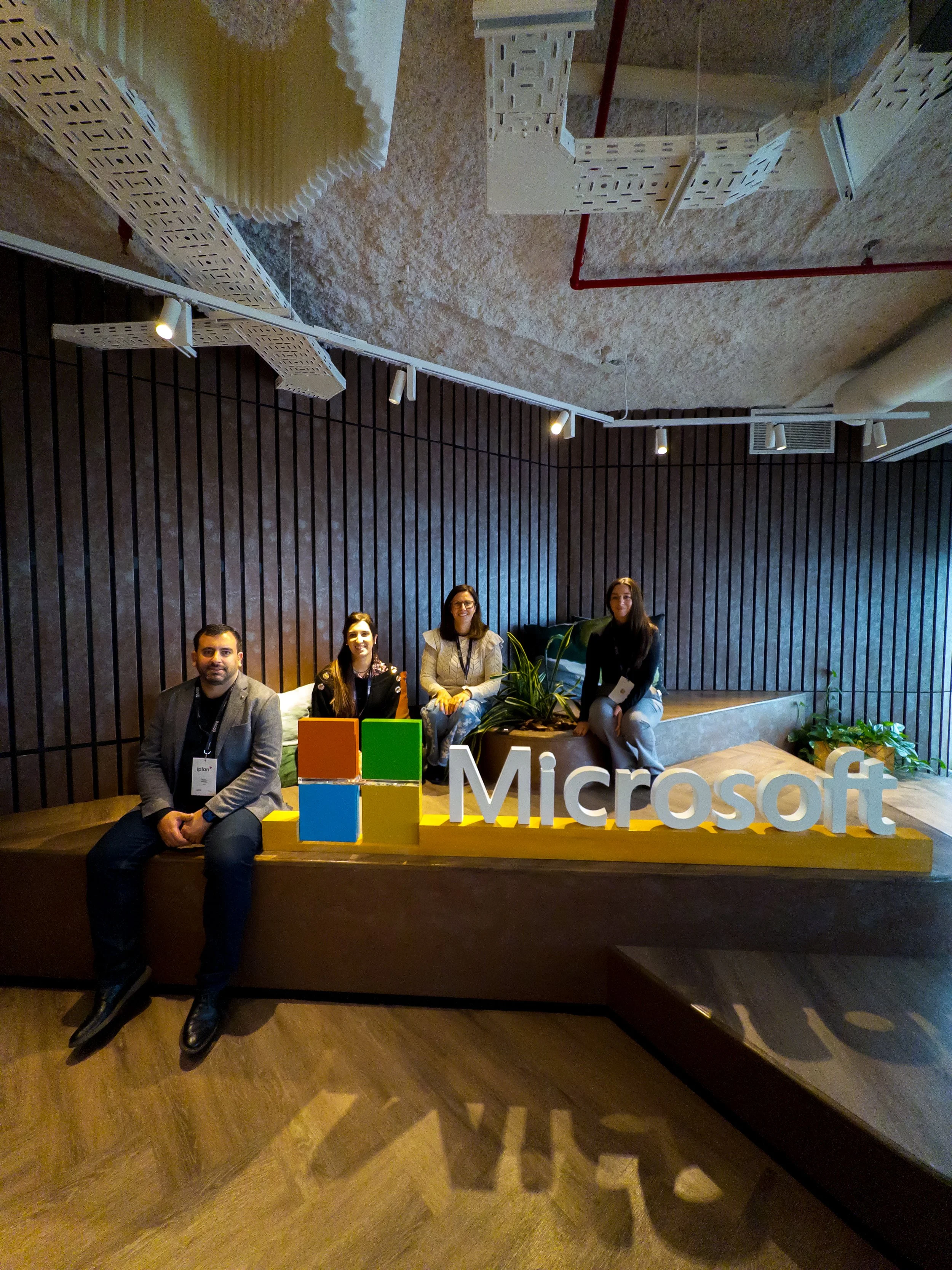 Cuatro personas sentadas frente a un cartel de Microsoft con el logotipo de cuatro bloques de colores en la parte inferior. La escena parece ser en un evento o conferencias, y el fondo tiene paredes con paneles de madera y una planta en el centro.