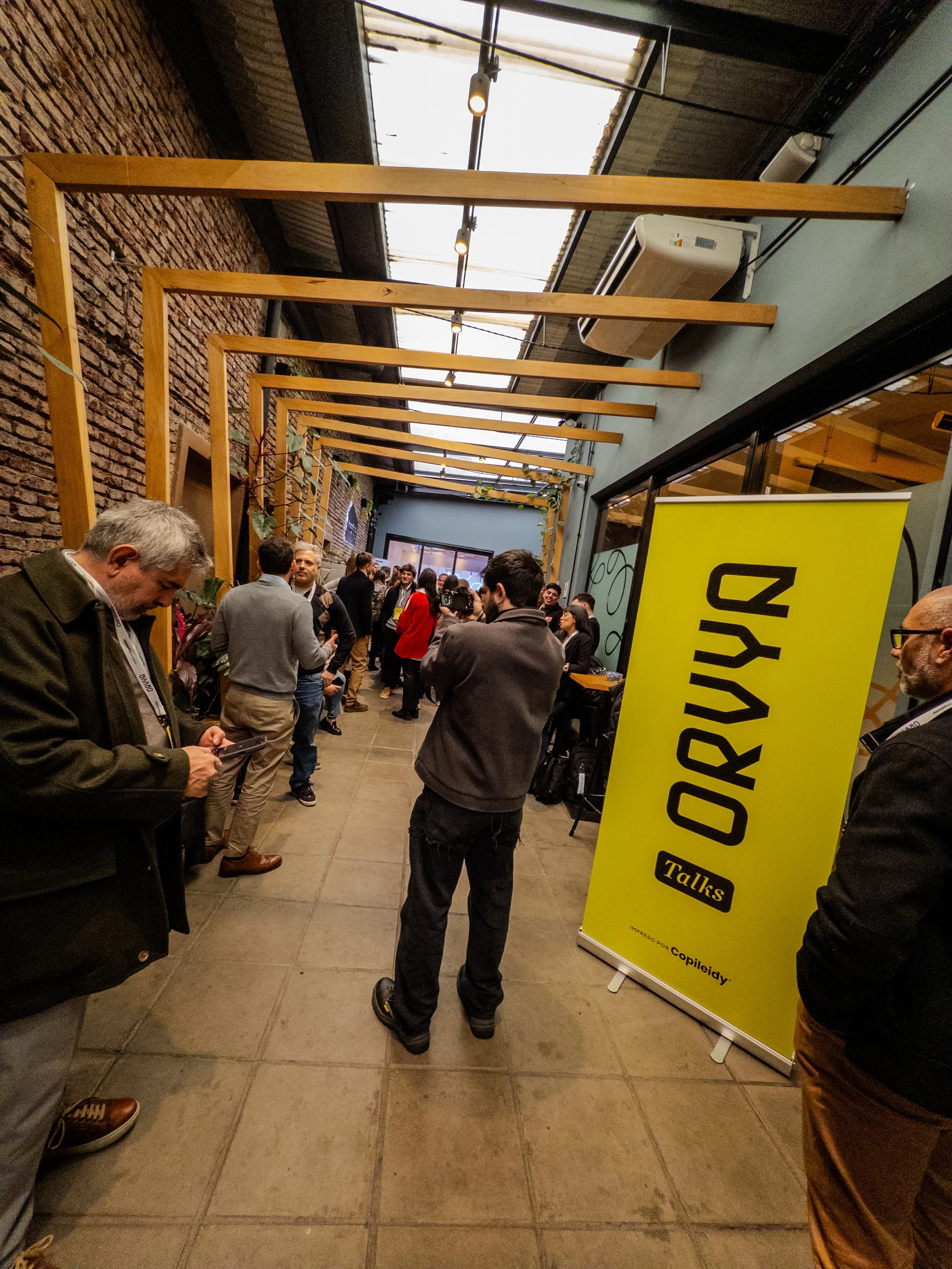 Reunión o evento en interiores con varias personas conversando y una muestra de banner amarillo con texto en inglés y en español.