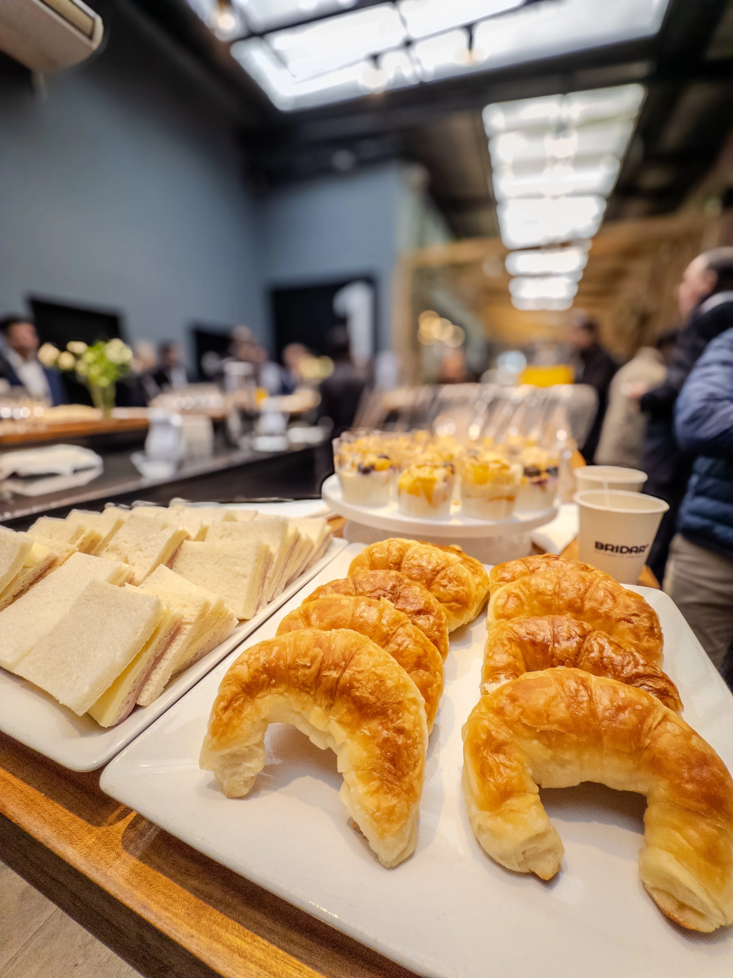 Una bandeja con sándwiches y croissants, además de vasos con líquidos y postres pequeños, en un entorno de reunión o celebración.