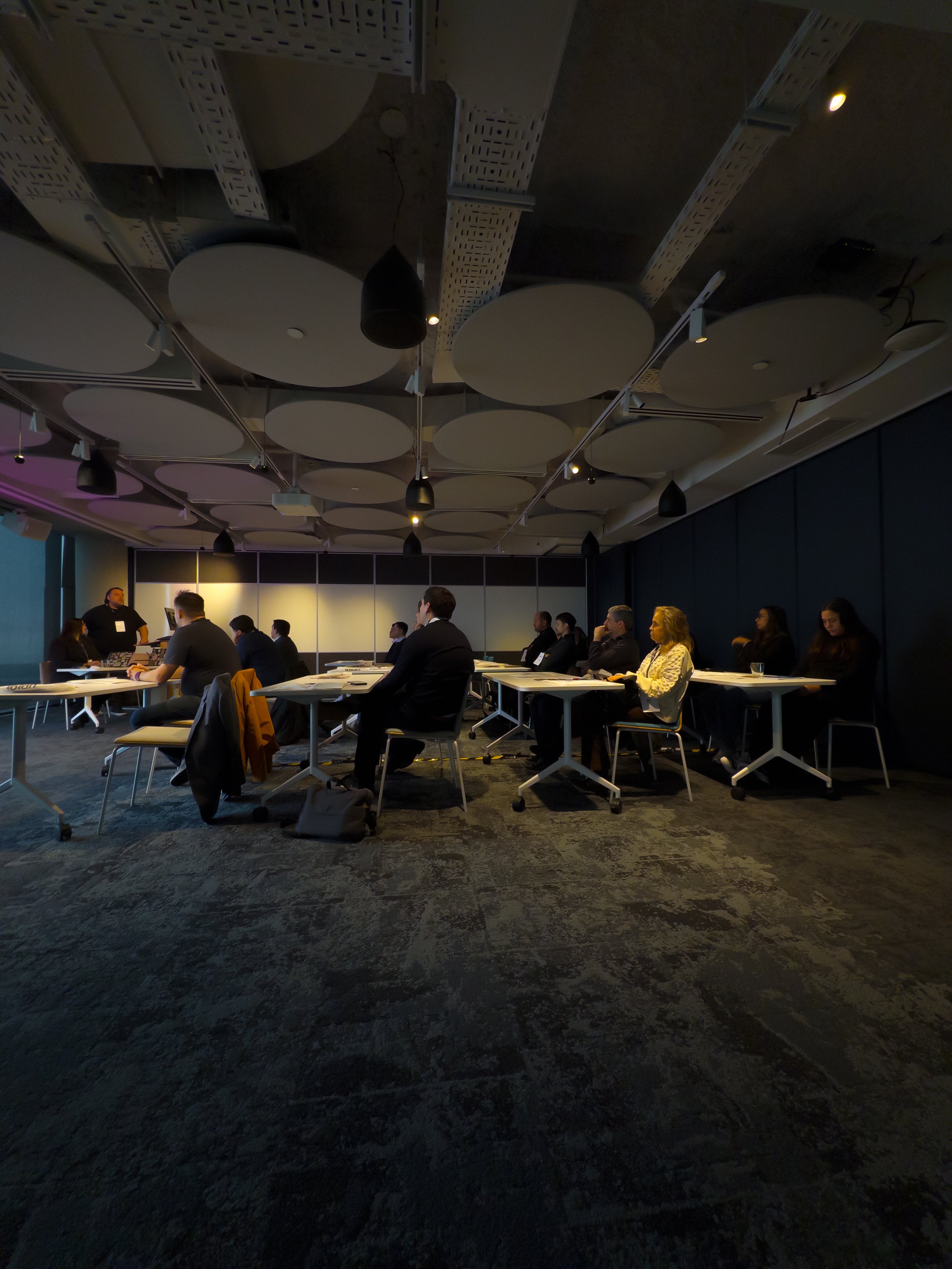 Reunión de personas en un salón moderno con techo acústico y luz tenue, sentados en mesas, en un entorno profesional.