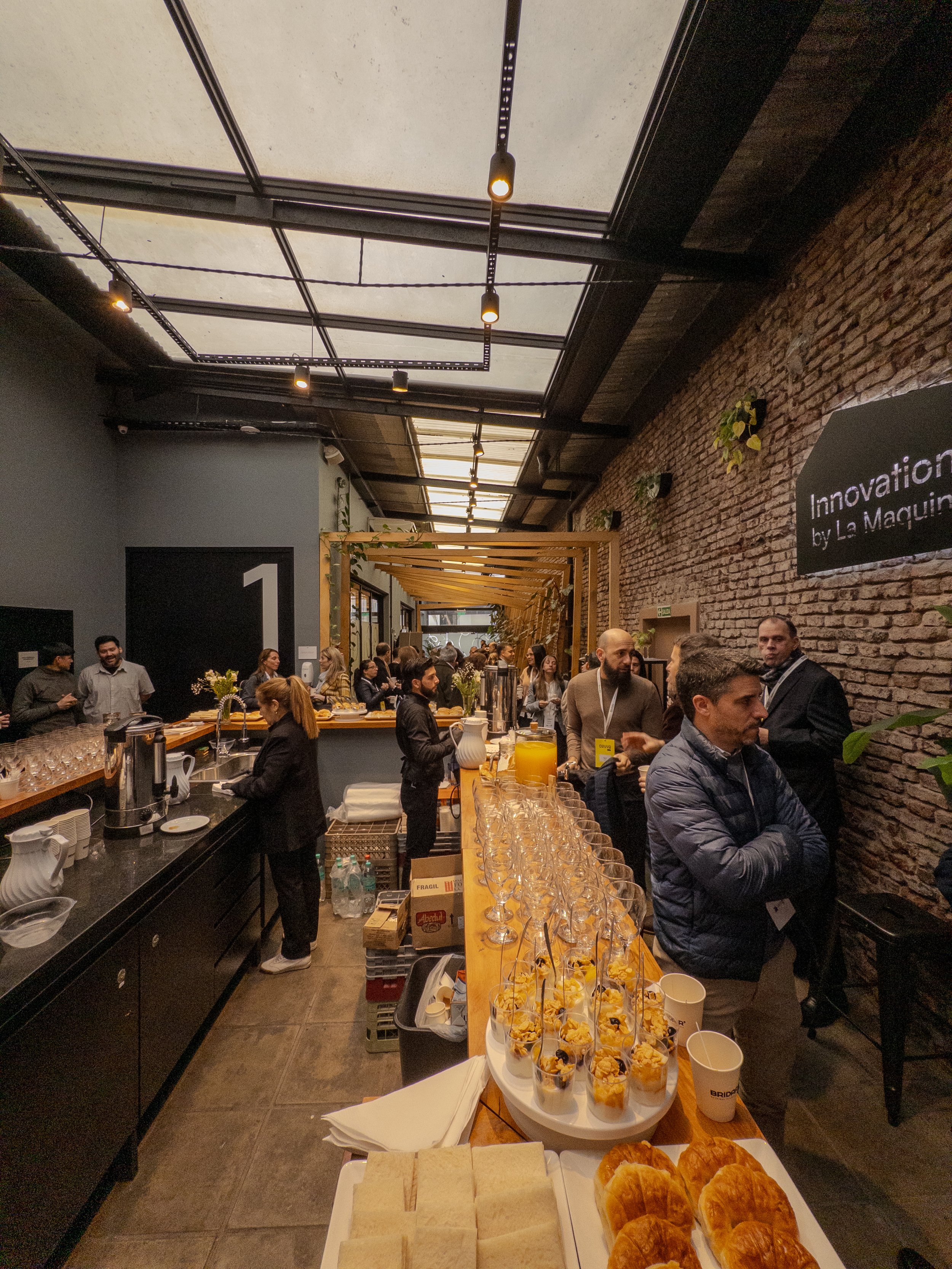 Una escena en un evento social con muchas personas en un espacio con paredes de ladrillo y un techo transparente que deja pasar la luz. En primer plano, hay vasos con comida y una bandeja con croissants. Al fondo, hay una barra con hielo y copas, y v
