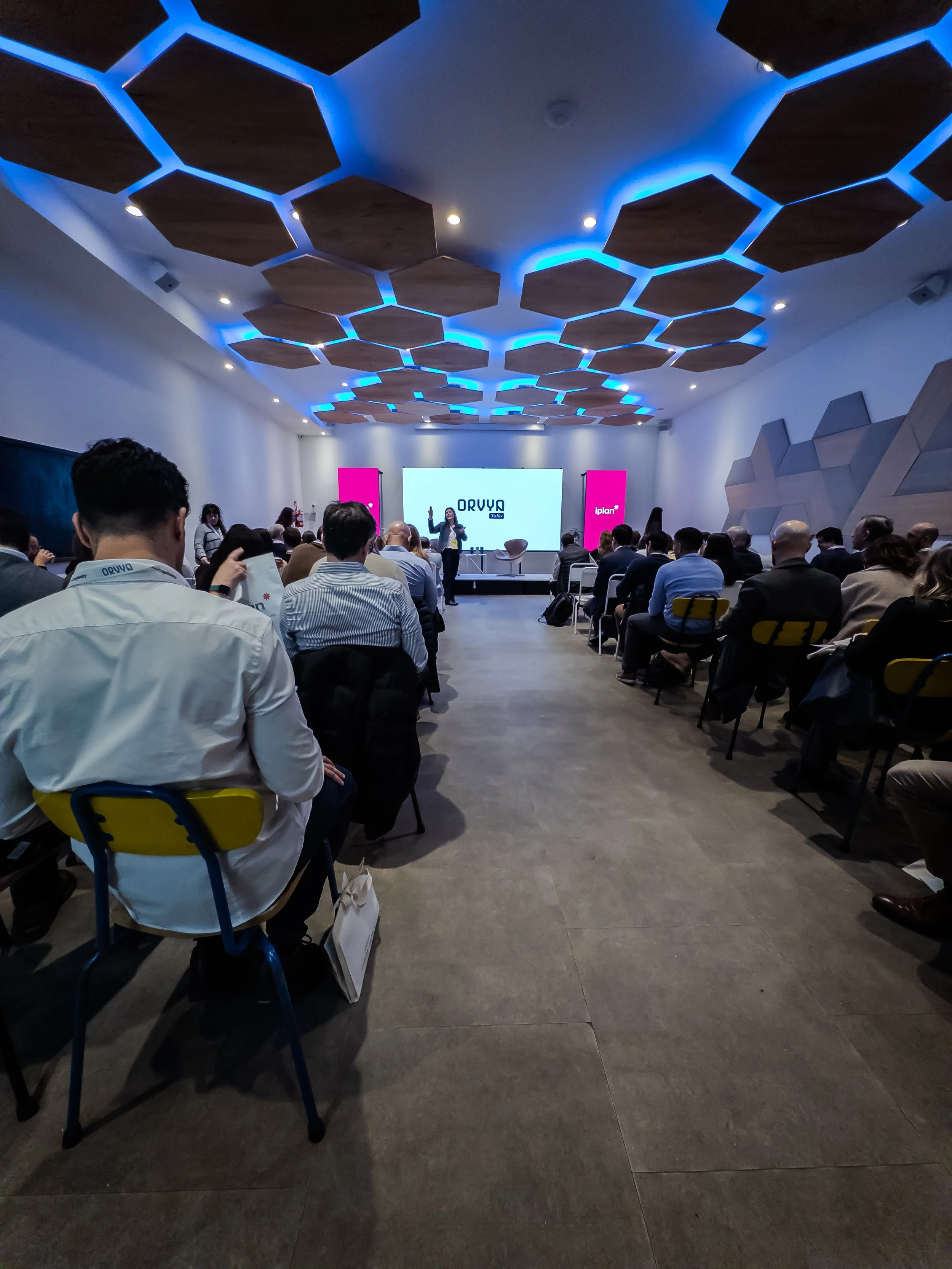 Escenario con una presentadora frente a una audiencia en una sala moderna con diseño en azulejos hexagonales de madera en el techo y luces LED azules. En el centro, pantalla grande con logotipo y dos paneles laterales rosas.